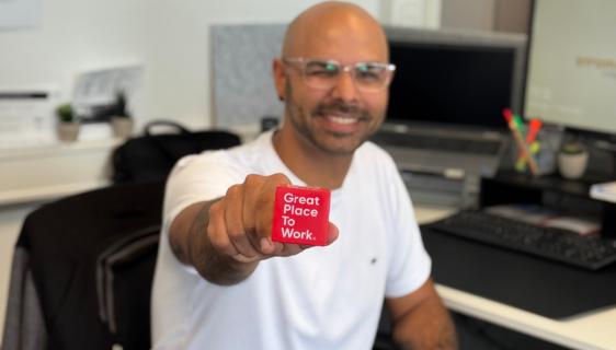 Mann im Büro lächelt in die Kamera und hält einen Würfel mit der Auffschrift "Great Place to Work" in der Hand.